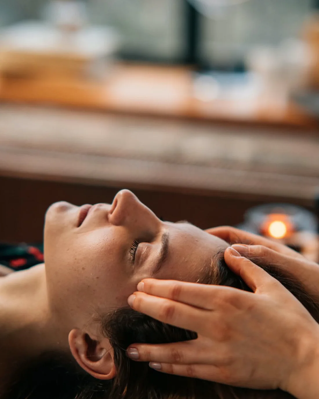 A practitioner's hands cradle the head of a woman resting with her eyes closed, a candle glowing softly behind